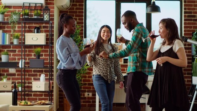 BIPOC mix of guests at home party sharing dance moves in living room, holding wine glasses in hand. Best friends dancing on energetic beats during weekend hangout, shaking hips