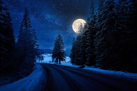 asphalt country road through forested countryside in winter at night. trees and hills in snow. mysterious atmosphere in full moon light