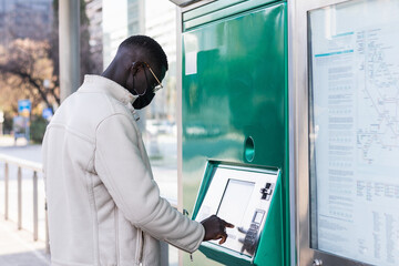 Black man buying a ticket to travel on public transport. He is wearing a medical mask as a...