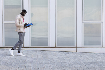 Black businessman in casual shirt walking near office building from outside, man smiling and using phone, freelancer typing message and browsing internet pages.