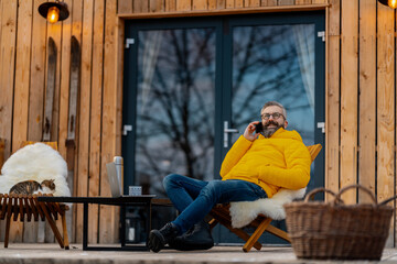 Mature man working from cozy cabin in mountains, sitting on terrace with laptop, enjoying cup of...