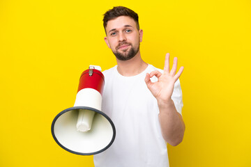 Young caucasian man isolated on yellow background holding a megaphone and showing ok sign with...