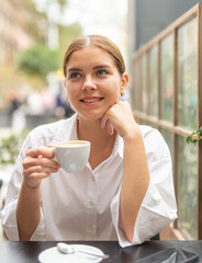 Smiling stylish young woman in white blouse drinking coffee sitting at the cafe