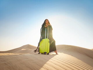 Arabian woman in traditional abaya costume with bright light green or yellow suitcase in sands....