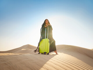 Arabian woman in traditional abaya costume with bright light green or yellow suitcase in sands. Concept and idea of travel to United Arab Emirates, sand dunes in desert of Dubai at sunset.