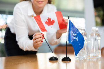 Female secretary places flags of the NATO (OTAN) and Canada on the table before the negotiations of top political figures - obrazy, fototapety, plakaty