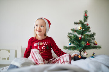 Happy preschooler girl wearing Christmas pajamas and playing with Christmas decorations on bed