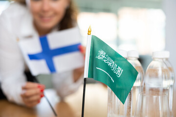 Little flag of Saudi Arabia on table with bottles of water and flag of Finland put next to it by positive young woman 