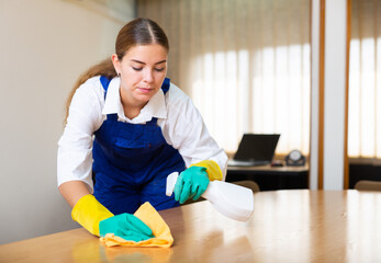 Skillful young workwoman of cleaning service wearing uniform and rubber gloves wiping dust on...