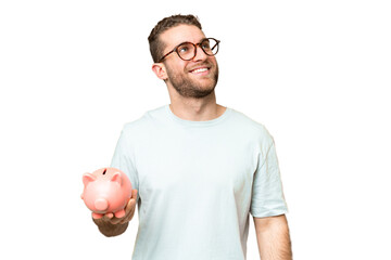 Young man holding a piggybank over isolated chroma key background looking up while smiling