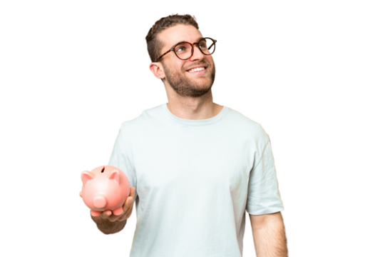 Young man holding a piggybank over isolated chroma key background looking up while smiling - Powered by Adobe
