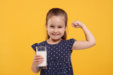 Cute girl with glass of fresh milk showing her strength on orange background