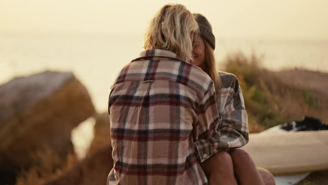A blonde girl in a black hat and a checkered shirt sits on the lap of her blonde boyfriend in a checkered shirt on the seashore Sunny morning at sunrise