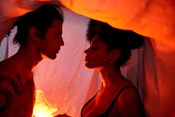 tempting diverse couple sitting under bedsheet and posing surrounded by lights, togetherness