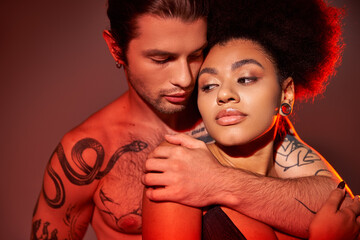 good looking tattooed man hugging warmly his african american girlfriend from behind, sexy couple