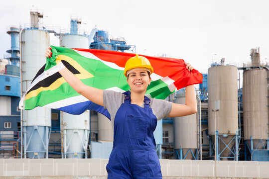 Cheerful Female Worker In Hardhat With South African Flag Standing In Front Of Factory