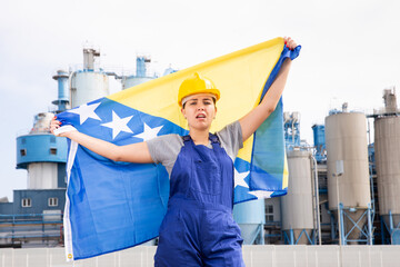 Sad unhappy young female worker in helmet waving national flag of Bosnia and Herzegovina during strike in front of big tanks at refinery factory
