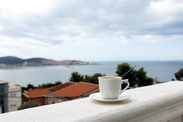 A white mug with coffee on a saucer stands on the balcony railing of the hotel room. Sea view from the apartment balcony