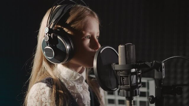 Girl in headphones sings in recording studio. surrounded by soundproof walls. Child demonstrates talent in front of microphone equipped with sound filter