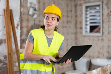 Focused young female architect in yellow protective workwear using modern software to create rooms design while standing in house under reconstruction