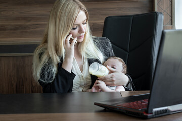 Young business woman feeds baby from bottle in office and talks on phone, concept of combining...