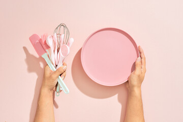 Baking flat lay. Female hands holding plate, kitchen tools, sieve, rolling pin, spatula and bruch on pastel pink background