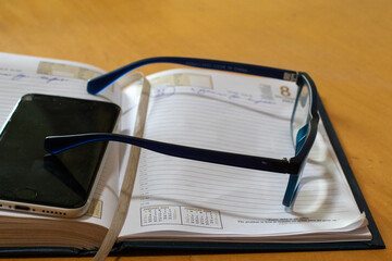 Reading glasses, a hard copy diary and a cell phone on a desk - obrazy, fototapety, plakaty