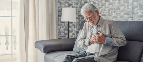 Worried elder senior man feeling bad, upset old middle aged grandfather touching chest feel sudden pain heartburn having heart attack sit on sofa at home.