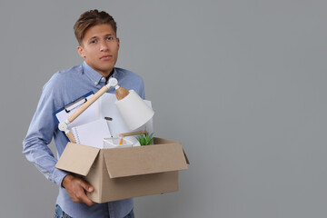Unemployed young man with box of personal office belongings on grey background. Space for text