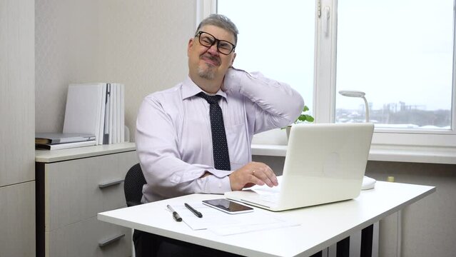The man is tired from working at the computer and is rubbing his stiff neck. The video is suitable for content about office work, workplace ergonomics and maintaining employee health.
