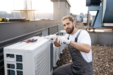 Focused builder worker dressed in grey overalls sitting and repairs air conditioner using...