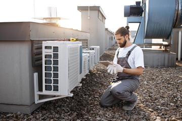 Serious repairman with braided hair crouching by air conditioner and looking at modern tablet screen outdoor. Caucasian skilled male working with gadget and searching for information on roof of plant. - Powered by Adobe