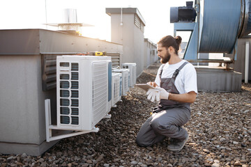 Serious repairman with braided hair crouching by air conditioner and looking at modern tablet screen outdoor. Caucasian skilled male working with gadget and searching for information on roof of plant. - Powered by Adobe