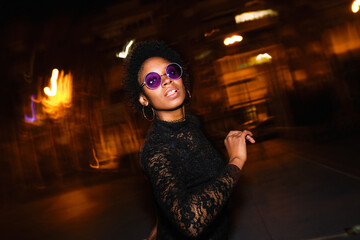 Confident Black woman in lace sweater and purple shades poses against a vibrant, blurry city night...