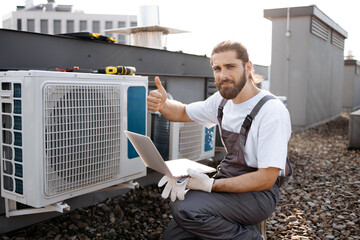 Bearded craftsman in uniform holding digital laptop while checking operation of fan in air conditioner with hand. Caucasian master crouching and using online manual for device outdoors, thumb up. - obrazy, fototapety, plakaty