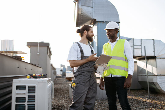 Skillful worker in overalls holding digital tablet while showing supervisor information found. Multicultural colleagues working with air conditioners and agreeing on further decisions on rooftop.