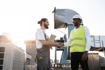 Smiling bearded worker in overalls standing and holding modern tablet while shaking hands with...