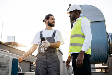 Serious craftsman in gray overall standing and looking at engineer in white hardhat while using...