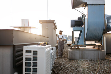 Concentrated bearded man wearing uniform holding toolkit and using digital tablet on factory. Caucasian professional craftsman walking by air conditioners and inspecting area on roof.