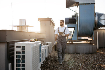 Concentrated bearded man wearing uniform holding toolkit and using digital tablet on factory. Caucasian professional craftsman walking by air conditioners and inspecting area on roof.