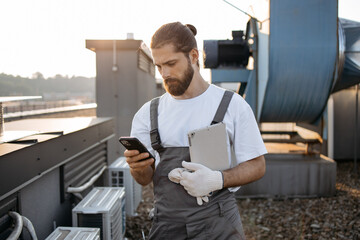 Focused bearded master dressed in overalls standing and holding tablet while using digital smartphone on fresh air. Caucasian skilled man using gadgets and maintaining equipment on roof of factory. - obrazy, fototapety, plakaty