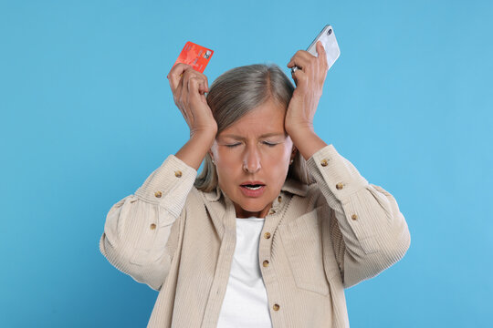 Upset woman with credit card and smartphone became a fraud victim on light blue background