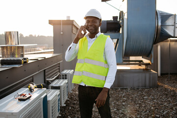 African american serious engineer wearing white helmet standing and having conversation on modern smartphone. Adult man performing work and resolving important issues with client on roof of factory.