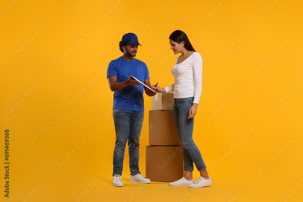 Poster Happy young woman signing order receipt on orange background. Courier delivery