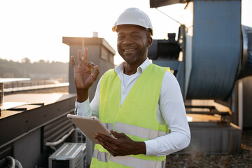 Serious factory worker standing and holding modern tablet in hands while servicing devices at...