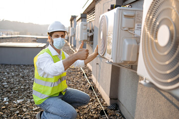 Professional worker of plant wearing dust mask crouching opposite air conditioner and providing...