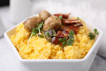 Cooked cornmeal with bacon, mushrooms and microgreens in bowl on white table, closeup