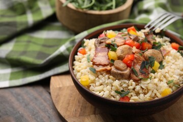 Cooked bulgur with vegetables, fried bacon and mushrooms in bowl on wooden table, closeup. Space for text