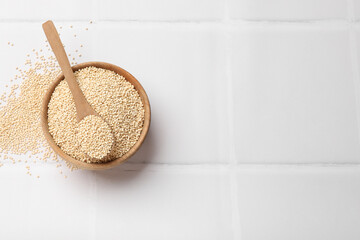 Dry quinoa seeds and spoon in bowl on white tiled table, top view. Space for text