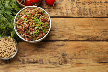 Delicious lentils with bacon, green onion in bowl, tomatoes and greens on wooden table, flat lay. Space for text